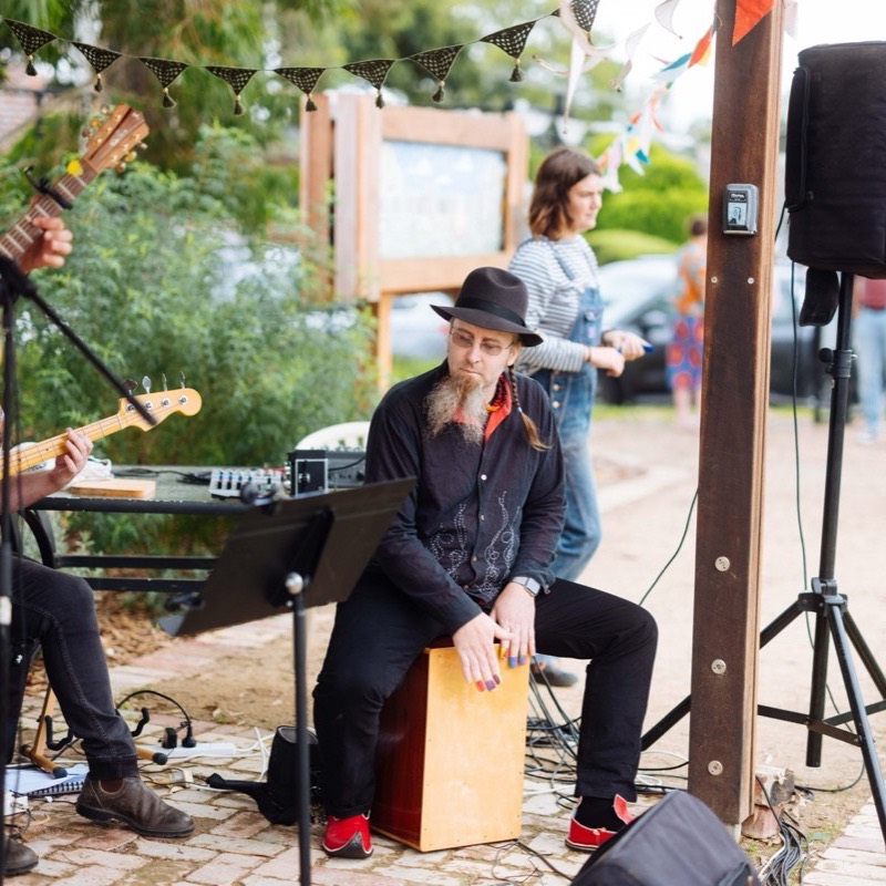 A guitarist and a cajon player performing beside the garden beds under strings of bunting