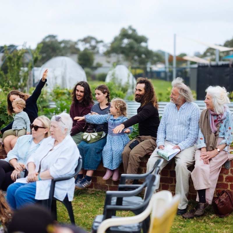 Adults and children sitting along a brick garden wall watching the music, one child pointing up