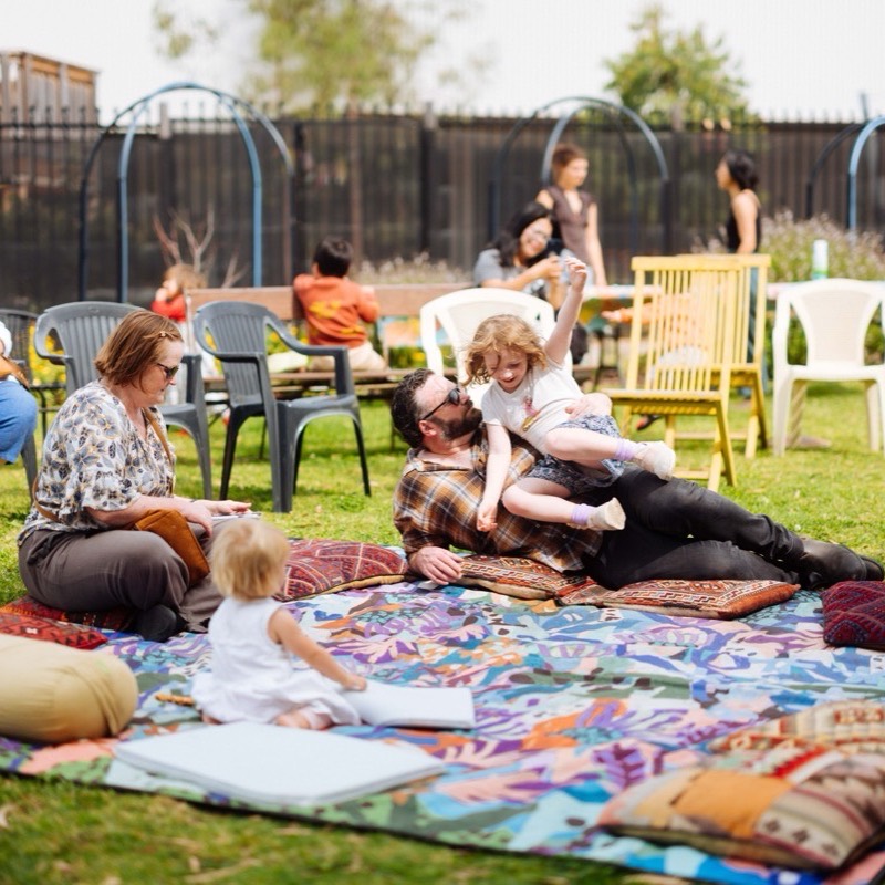 A parent lying on a picnic rug playing with a laughing child